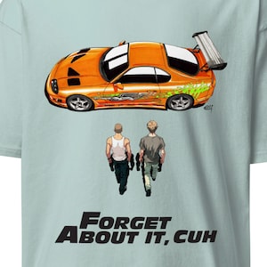 May include: Light blue t-shirt featuring an orange sports car graphic with green accents and two figures walking below. The text "FORGET ABOUT IT, CUH" is printed in black. The shirt has a crew neck and short sleeves.