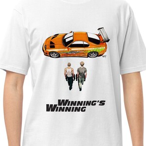 May include: White t-shirt featuring an orange sports car graphic with a rear spoiler and green accents. Below the car are two figures walking away, with the text "Winning's Winning" in bold black letters.
