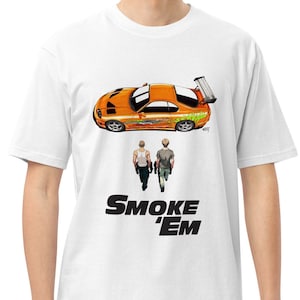 May include: White t-shirt featuring an orange sports car graphic with green accents and two figures walking below. The text "SMOKE 'EM" is printed in black. The shirt is made of a soft, comfortable material.