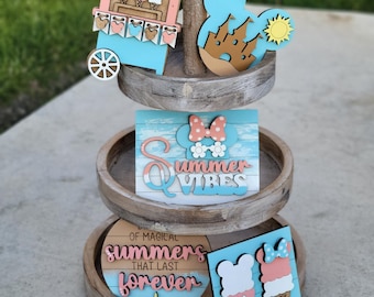 May include: A three-tiered wooden tiered tray decorated with various Disney-themed wooden cutouts. The top tier features a blue and pink ice cream stand with the words "ICE CREAM" above it. The middle tier features a blue and white sign with the words "Summer Vibes" and a pink and white Minnie Mouse head. The bottom tier features a blue and white sign with the words "Of Magical Summers That Last Forever" and two ice cream cones with pink and white Mickey Mouse ears.