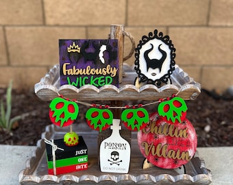 May include: A two-tiered wooden tiered tray with a Halloween theme. The top tier features a purple and black sign that says "Fabulously Wicked" with a crown and silhouette of a woman. The bottom tier features a green and red apple poison sign, a white sign that says "Poison Do Not Drink", a red and white sign that says "Chillin Like A Villain", and a stack of three black, green, and red blocks that say "Just One Bite".