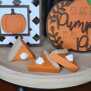 May include: A wooden tray holds three orange pumpkin pie slices topped with white whipped cream. A decorative pumpkin sign and a pumpkin-themed framed picture are in the background. The overall theme is autumn and fall.
