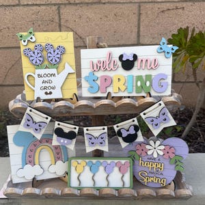 May include: A tiered wooden stand with various spring-themed wooden signs and decorations. The signs feature Mickey Mouse ears, butterflies, rainbows, and the words "Welcome Spring", "Bloom and Grow", and "Have a Happy Magical Spring".
