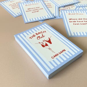 May include: A card game titled "The Bach Club" with a light blue and white striped design. The cover shows red lingerie and the words "CARD GAME". Several individual cards with questions are scattered around the deck.