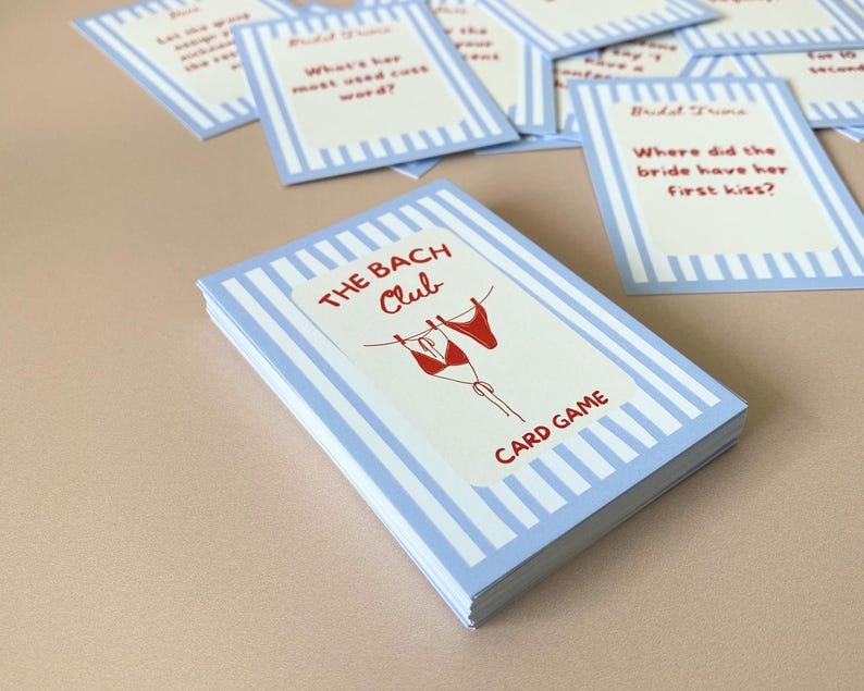 May include: A card game titled "The Bach Club" with a light blue and white striped design. The cover shows red lingerie and the words "CARD GAME". Several individual cards with questions are scattered around the deck.