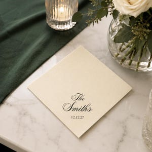 May include: A square cream-colored paper napkin featuring black printed text that reads The Smiths followed by the date 12.12.27 in a classic serif font. The napkin is positioned flat on a surface, displaying a smooth, matte texture.