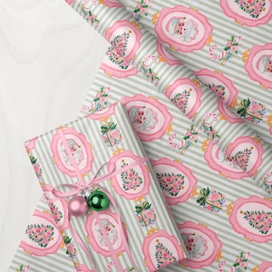 May include: Wrapping paper featuring a repeating pattern of Santa Claus faces and floral Christmas trees inside pink scalloped frames on a light gray and white striped background, with a gift wrapped in the same paper and tied with a pink ribbon.