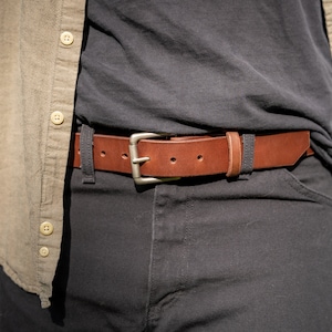 May include: A brown leather belt with a silver buckle. The belt is worn around the waist of a person wearing a black shirt and black pants.