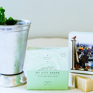 Könnte beinhalten: Zwei Schachteln handgemachter Seife mit Minz-Julep-Duft. Die Schachteln sind grün und weiß mit dem Text "My City Soaps" und "Highland Mint Julep". Die Seife hat die Form eines Blocks und ist hellgrün. Im Hintergrund steht ein silberner Becher mit einem Minzblatt.
