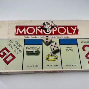 May include: A vintage Monopoly board game box featuring the iconic top-hatted mascot. The board surface displays the GO space, Boardwalk, Luxury Tax, Park Place, and a Chance square. Two cream-colored dice and a silver car token are included.