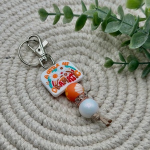 May include: A white keychain with a rainbow design and the text "A little bit dramatic". The keychain has a silver metal ring and a white bead with a silver accent.