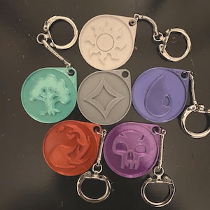 May include: Set of seven keychains featuring different colored, 3D printed symbols. The symbols include a sun, a tree, a diamond, a teardrop, a flame, a skull, and a circle.