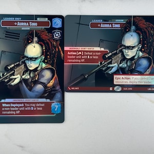 May include: Two trading cards featuring the character Aurra Sing, a leader unit and assassin. The cards have a dark background with a portrait of a pale-skinned character with red dreadlocks aiming a rifle. Text includes "Leader Unit" and "Underworld - Bounty Hunter."