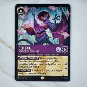 May include: A collectible card featuring the character Demona from the Wyvern Clan. The card has a dark background with an illustration of Demona, a purple-skinned gargoyle, holding a book. Text includes character abilities and game details.