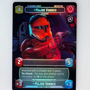 Puede incluir: Una carta de juego de cartas coleccionables con Major Vonreg, un personaje de Red Baron del universo de Star Wars. La carta es negra con acentos holográficos y presenta una ilustración detallada de Major Vonreg con su armadura roja. El texto de la carta dice "Al atacar: Puedes dar a otra unidad en esta arena +1/+0 para esta fase."