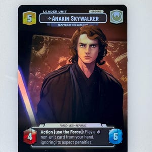 May include: Anakin Skywalker trading card. The card features a portrait of Anakin Skywalker, with the text "TEMPTED BY THE DARK SIDE." The card has a black border and includes game information such as "LEADER UNIT" and "GROUND."