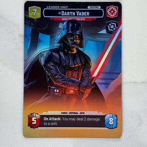Puede incluir: Carta coleccionable de Star Wars con Darth Vader, el Señor Oscuro de los Sith, sosteniendo un sable de luz rojo. La tarjeta tiene un borde negro y el texto "LEADER UNIT" y "DARTH VADER". También muestra los números 7, 5 y 8.