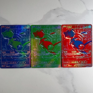 Puede incluir: Tres cartas coleccionables rectangulares con el personaje Mew, cada una con un esquema de color holográfico distinto en azul, verde y rojo. Las cartas muestran el nombre Mew, 60 HP, el tipo Psíquico y texto del juego con acabado metálico.