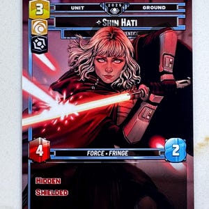May include: A trading card featuring Shin Hati, with a red lightsaber and a hooded cloak. The card has a black background with the text "FORCE FRINGE" and "HIDDEN SHIELDED". The card also has the number "3", "4", and "2".