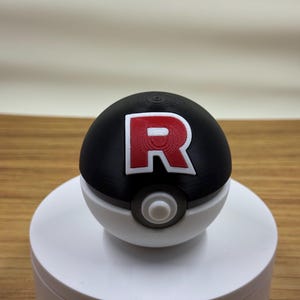 Puede incluir: Réplica impresa en 3D de una Poké Ball. La mitad superior es negra con un emblema "R" rojo y blanco, la mitad inferior es blanca y tiene un botón central gris. Se muestra sobre un soporte circular blanco, sobre un fondo neutro.
