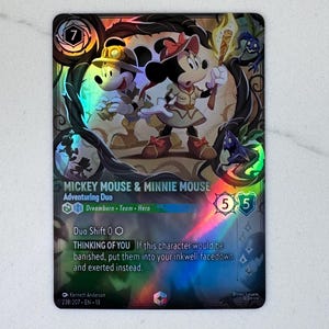 Könnte beinhalten: Eine holografische Disney Lorcana Sammelkarte mit Mickey Mouse und Minnie Mouse als Abenteurer-Duo. Die Karte zeigt den Text Mickey Mouse & Minnie Mouse, Adventuring Duo und die Fähigkeit Thinking of You mit Kosten von 7.