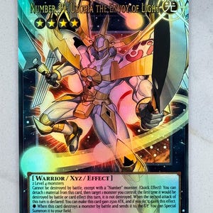Puede incluir: Carta coleccionable holográfica con "Número 39: Utopía, el Enviado de la Luz". La carta muestra un guerrero con armadura dorada y púrpura, empuñando una espada y un escudo. El texto detalla sus atributos y habilidades, con ATK 2500 y DEF 2000.