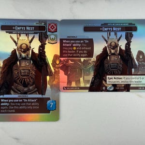 May include: Two collectible game cards featuring the character "Enfys Nest." The cards have a futuristic design with a central figure in a helmet and armor. Text on the cards describes abilities and game mechanics. The cards are primarily blue, black, and gold.