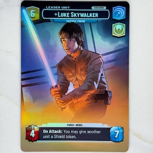 May include: A Star Wars trading card featuring Luke Skywalker, holding a blue and pink lightsaber. The card has a blue and gold border with the text "LEADER UNIT" and "LUKE SKYWALKER". The card also has the text "FORCE - REBEL".