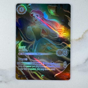 May include: A holographic trading card featuring Ariel from Disney's The Little Mermaid. The card has a shimmering effect with a portrait of Ariel in a flowing gown. Text includes "ARIEL Ethereal Voice" and "COMMAND PERFORMANCE".