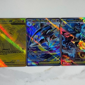 Puede incluir: Tres cartas coleccionables rectangulares con Mega Charizard X ex y Mega Sharpedo EX. La carta de la izquierda es dorada con acabado holográfico, la central muestra una criatura tiburón azul y la de la derecha un dragón oscuro con fuego.
