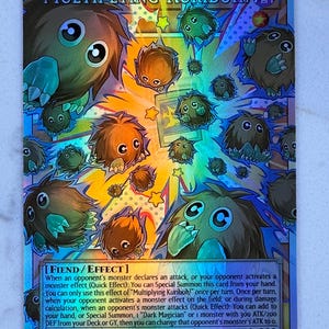 May include: A holographic trading card featuring the text "Multiplying Kuriboh!" and various cartoonish creatures. The card has a dark background with a colorful design and the text "FIEND/EFFECT". The card's ATK is 300 and DEF is 200.