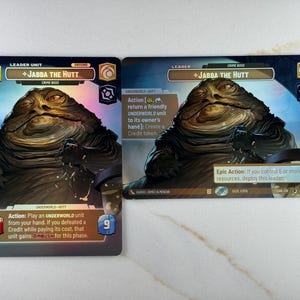 May include: Two Star Wars trading cards featuring Jabba the Hutt. The cards have a brown and blue color scheme with text that reads "Leader Unit" and "Crime Boss". The cards also have action descriptions and numerical values.