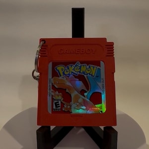 May include: An orange Game Boy cartridge with the Pokémon logo and a Charizard illustration. The cartridge has a silver key ring attached. The text "GAME BOY" is printed at the top.