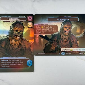 May include: Two trading cards featuring Chewbacca, a character from the Star Wars franchise. The cards have a brown and tan color scheme with text that reads "Leader Unit" and "Hero of Kessel". One card has an "On Attack" description.