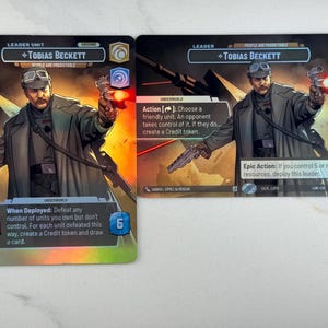 Puede incluir: Dos cartas coleccionables con el personaje Tobias Beckett. Las cartas muestran a un hombre con un abrigo largo y gafas sosteniendo un blaster. Las cartas tienen diferentes textos y gráficos, incluyendo las palabras "Leader Unit" y "Underworld."