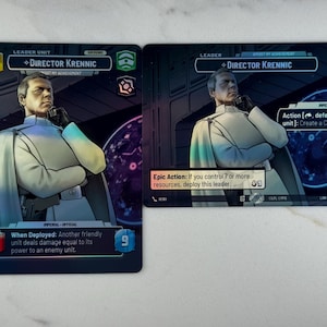 Puede incluir: Dos cartas coleccionables con el personaje Director Krennic de Star Wars. Las cartas tienen un fondo oscuro con un retrato del personaje. Las cartas incluyen texto que describe las habilidades y estadísticas del personaje. Las cartas son rectangulares.