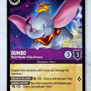 May include: A Dumbo trading card featuring the character with large pink ears, a yellow hat, and a red collar. The card includes the text "DUMBO Ninth Wonder of the Universe Storyborn Hero". The card has a purple and blue background.