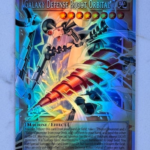 May include: A holographic trading card featuring "Galaxy Defense Robot Orbital 7." The card depicts a mechanical robot with a red drill-like weapon, set against a backdrop of blue and gold energy. The card's text details its effects and stats.