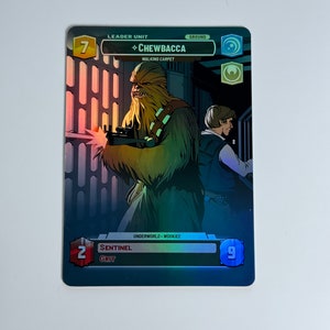 May include: A Star Wars collectible card featuring Chewbacca, a Wookiee character from the Star Wars franchise. The card is blue and green with a holographic finish. The card has a number 7 in the top left corner and the text "LEADER UNIT" and "CHEWBACCA" in the top center. The card also has the text "WALKING CARPET" and "GROUND" in the top right corner. The card has a number 2 in the bottom left corner and the text "SENTINEL" and "GRIT" in the bottom center. The card has a number 9 in the bottom right corner.