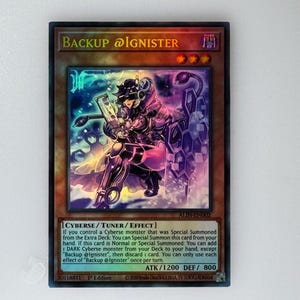 May include: A "Backup @Ignister" trading card featuring a purple and blue robot character with a glowing background. The card text reads "If you control a Cyberse monster that was Special Summoned from the Extra Deck: You can Special Summon this card from your hand. If this card is Normal or Special Summoned: You can add 1 DARK Cyberse monster from your Deck to your hand, except "Backup @Ignister", then discard 1 card. You can only use each effect of "Backup @Ignister" once per turn."
