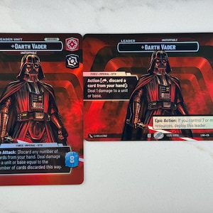 May include: Two Star Wars trading cards featuring Darth Vader. The cards have a red and black color scheme with the character's image prominently displayed. Text on the cards includes "Leader Unit", "Unstoppable", and game instructions. The cards are rectangular.
