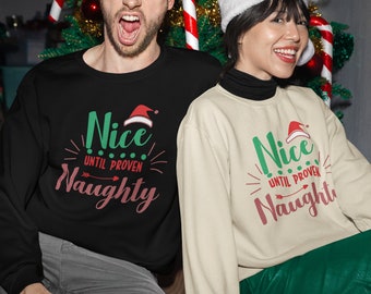 May include: Two people wearing matching Christmas jumpers with the text "Nice Until Proven Naughty" in green and pink. The jumpers feature a red Santa hat graphic. The people are sitting in front of a Christmas tree.