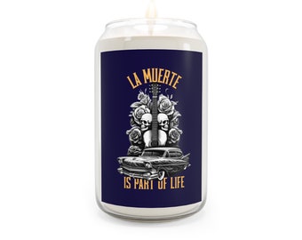 May include: A white candle in a glass jar with a blue label. The label features a black and white illustration of a car, two skulls, and a guitar with roses. The text on the label reads "La Muerte is part of life."
