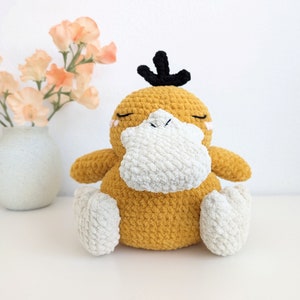 Puede incluir: Un juguete de peluche tejido a crochet con forma de pato, con cuerpo amarillo, pico blanco y ojos negros. El juguete está sentado sobre una superficie blanca.