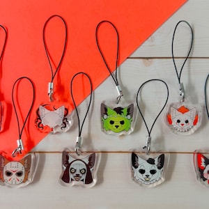 May include: A collection of clear acrylic charms featuring cartoon cat faces inspired by horror movie characters. Each charm has a black lanyard and silver clasp, set against a bright orange background and a white wood-grain surface.