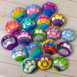 Pode incluir: Uma coleção de pelúcias redondas e coloridas, cada uma com um design de pegada em vários esquemas de cores de bandeiras do orgulho. As pelúcias são dispostas em uma superfície de madeira clara, mostrando uma variedade de bandeiras do orgulho LGBTQ+.