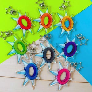 Może przedstawiać: Kolekcja kolorowych breloków akrylowych. Każdy brelok przedstawia literę "O" w innym kolorze, otoczoną wzorem rozbłysku gwiazdy. Breloki mają srebrne zapięcia w kształcie gwiazd.