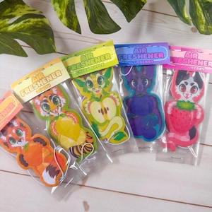 May include: A collection of air fresheners in clear plastic packaging. Each air freshener has a unique cartoon animal design and a corresponding fruit scent label. The scents include lemon, green apple, blueberry, and strawberry.