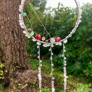 Pode incluir: Um apanhador de sonhos artesanal com um anel de metal prateado adornado com cristais rosa e transparentes. Contas coloridas e fios de cristal pendurados pendem do anel. O fundo apresenta uma árvore e vegetação.