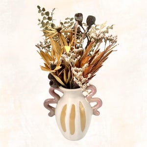 May include: A white ceramic vase with pink handles and yellow stripes. The vase is filled with a bouquet of dried flowers, including brown, beige, and white flowers.